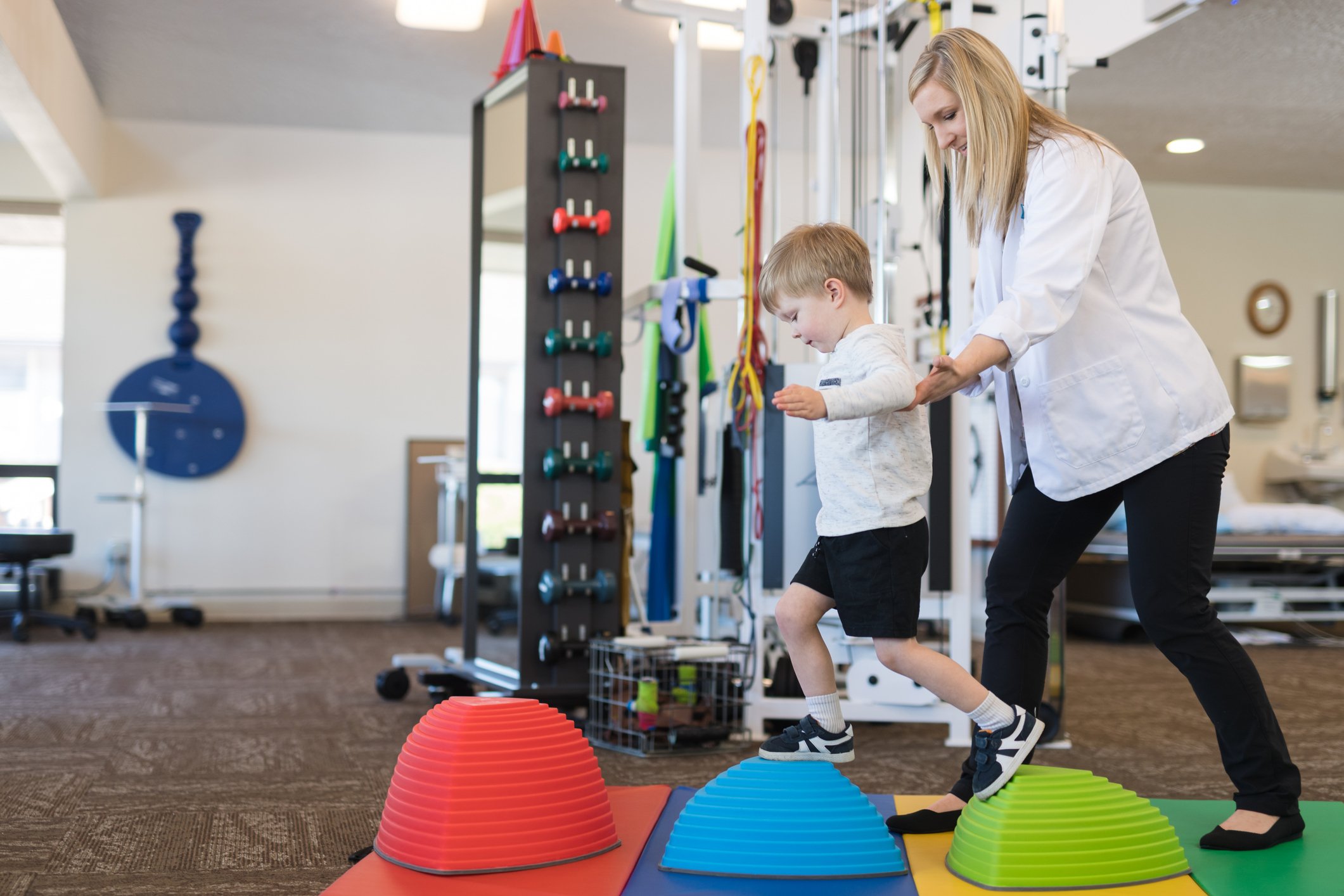 A blonde physical therapist works with a young boy in an open floor clinic. She is helping him do a fun exercise on a trio of colorful balance toys on the floor. She is gently guiding him from behind as he carefully steps from one to the next.