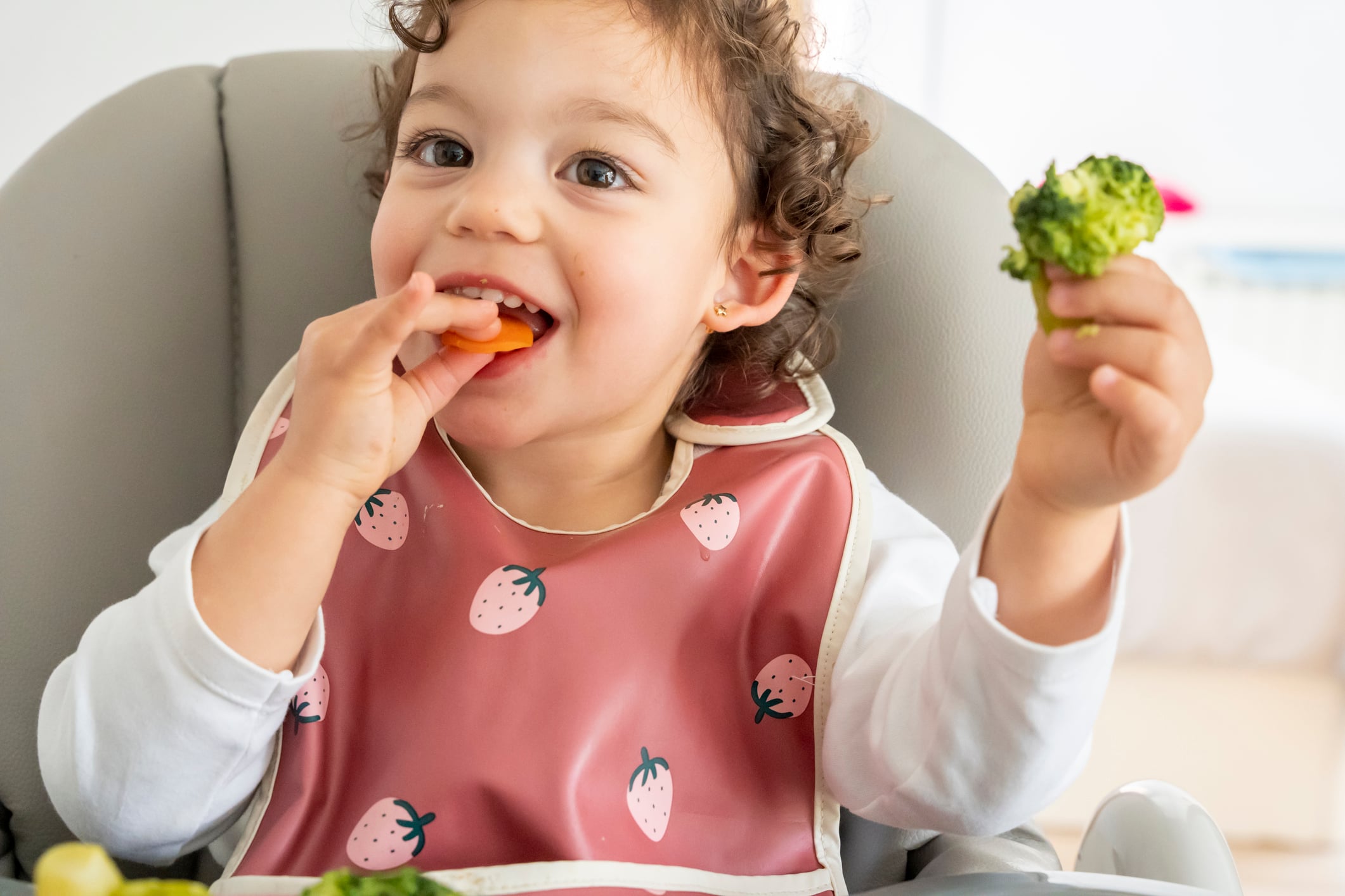 little girl sitting in her high chair enjoying eating vegetables, holding broccoli and carrot in her hand. healthy eating in childhood, care and well-being of children. baby led weaning.